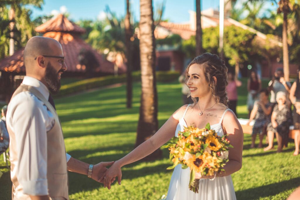 A joyful bride and groom hold hands during a romantic sunny outdoor wedding ceremony surrounded by lush greenery.